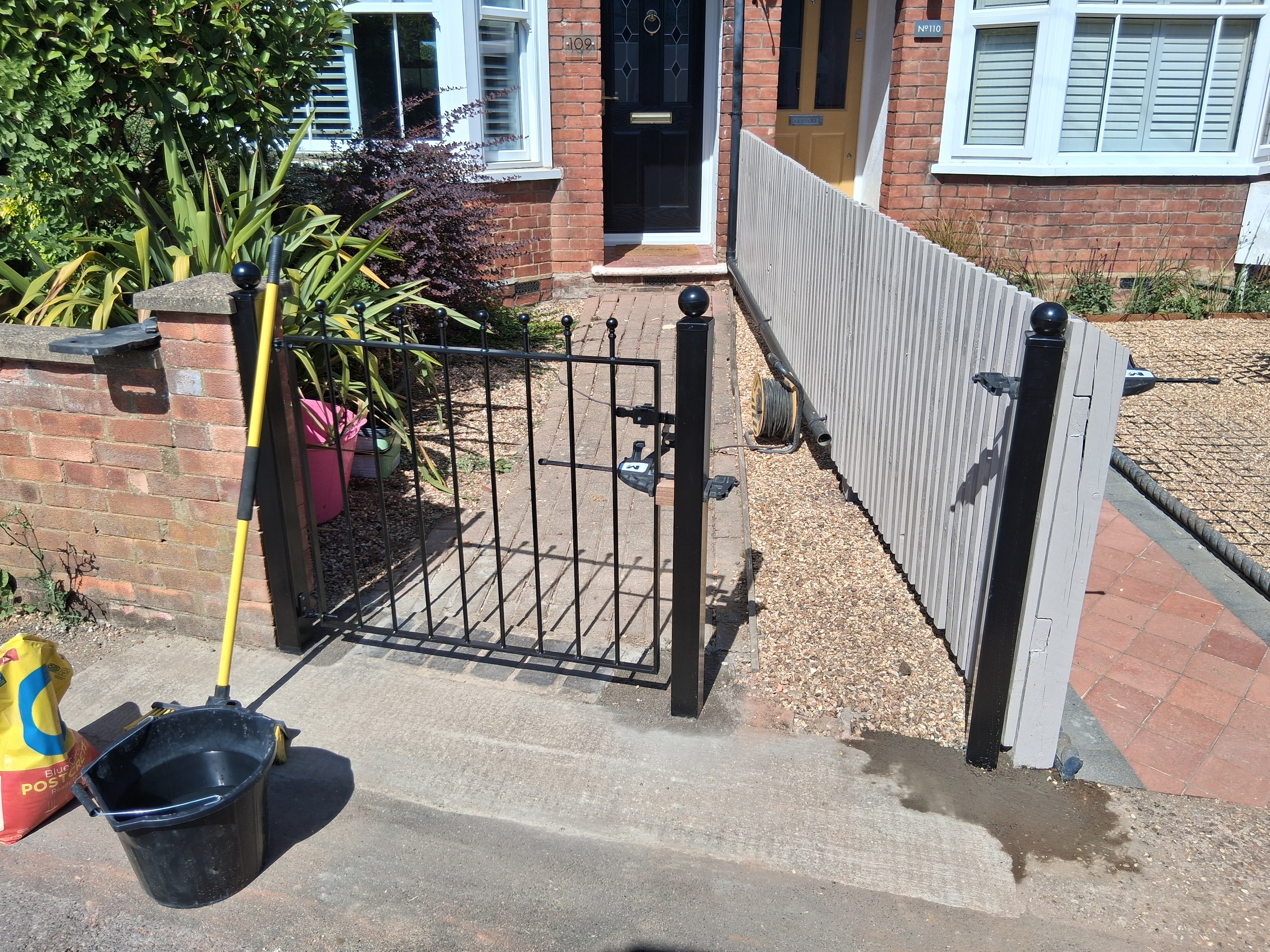 Black ball-top pedestrian gate hung on square metal posts at the head of a front path, with a timber fence panel alongside